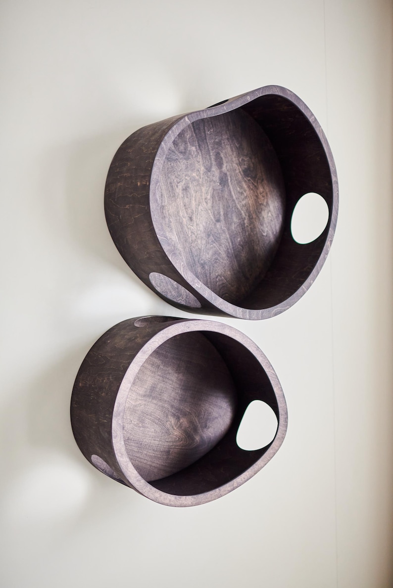 May include: Two dark brown wooden wall shelves with a unique, curved design. Each shelf has a circular opening on the side. The shelves are mounted on a light-colored wall, showcasing their modern aesthetic.