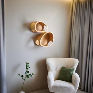 May include: Two wooden, organically shaped wall shelves with a light finish are mounted on a neutral wall. Below, a white, textured armchair with a green pillow sits near a vase with greenery. The room has a modern, minimalist aesthetic.