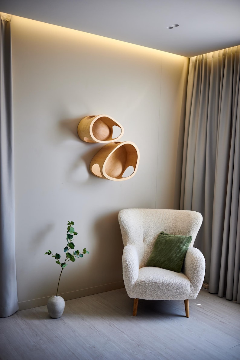 May include: Two wooden, organically shaped wall shelves with a light finish are mounted on a neutral wall. Below, a white, textured armchair with a green pillow sits near a vase with greenery. The room has a modern, minimalist aesthetic.