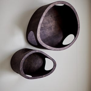 May include: Two dark brown, oval-shaped wall shelves with a smooth finish. Each shelf has a large opening and a smaller, teardrop-shaped cutout. The shelves are mounted on a light gray wall.