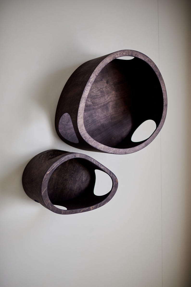 May include: Two dark brown, oval-shaped wall shelves with a smooth finish. Each shelf has a large opening and a smaller, teardrop-shaped cutout. The shelves are mounted on a light gray wall.
