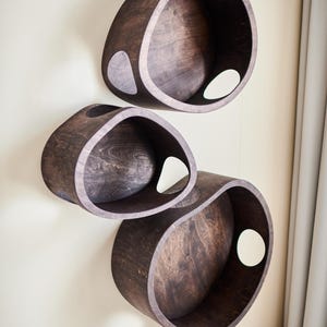 May include: Three dark brown, oval-shaped wooden shelves mounted on a light beige wall. Each shelf has a large, circular opening and a smaller, teardrop-shaped cutout. The shelves are arranged vertically.