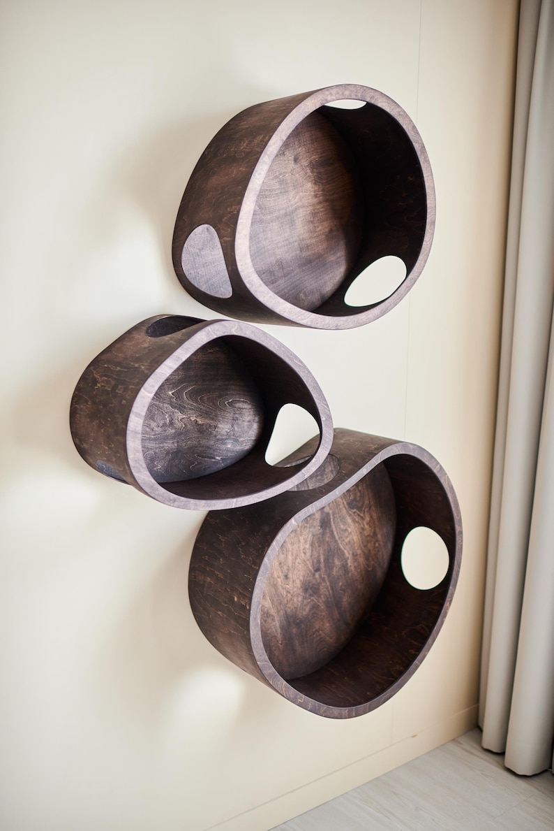 May include: Three dark brown, oval-shaped wooden shelves mounted on a light beige wall. Each shelf has a large, circular opening and a smaller, teardrop-shaped cutout. The shelves are arranged vertically.