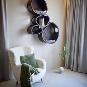 May include: A modern cat shelf unit with multiple oval-shaped compartments in dark wood, mounted on a wall. Each compartment has a gray cushion. A white armchair with a green pillow and blanket sits below the shelves.