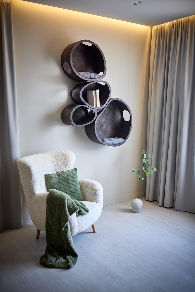 May include: A modern cat shelf unit with multiple oval-shaped compartments in dark wood, mounted on a wall. Each compartment has a gray cushion. A white armchair with a green pillow and blanket sits below the shelves.