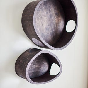 May include: Two dark brown wooden wall shelves with a unique, organic shape. Each shelf has a large, open interior and a circular cutout on the side. The shelves are mounted on a light-colored wall, creating a modern, minimalist aesthetic.