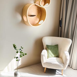 May include: A modern cat wall shelf in a light wood finish. The design features two connected, rounded structures with circular openings. A white armchair with a green pillow sits below. A vase with greenery is on the floor.