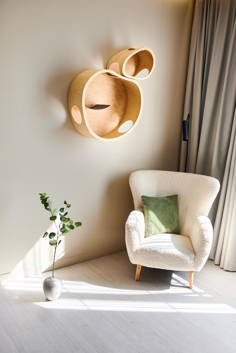 May include: A modern cat wall shelf in a light wood finish. The design features two connected, rounded structures with circular openings. A white armchair with a green pillow sits below. A vase with greenery is on the floor.
