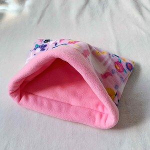 Könnte beinhalten: Ein rosa Haustierbett aus Fleece mit einem Blumen- und Pfotenabdruckmuster auf der Außenseite. Die Innenseite ist einfarbig rosa. Das Bett ist an einem Ende offen und schafft einen gemütlichen, geschlossenen Raum für kleine Haustiere.