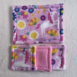 Può includere: Set di articoli in tessuto pile rosa con un motivo a unicorno e floreale. Il set include un grande rettangolo e tre quadrati più piccoli. L'unicorno ha una criniera e una coda rosa. I fiori sono gialli, rosa e blu.