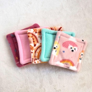 Op de afbeelding: Vier kleine, vierkante, met fleece gevoerde pads met verschillende kleurrijke patronen. De pads zijn roze, turquoise en oranje met verschillende ontwerpen, waaronder regenbogen, wolken en dieren.