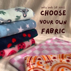 Può includere: Una pila di traversine in tessuto piegate con vari motivi tra cui koala, stelle bianche su blu, ciliegie rosse su blu navy, fragole su rosa, un motivo a scacchi rosa e un design arcobaleno arancione con il testo potty pads (all sizes) CHOOSE YOUR OWN FABRIC.