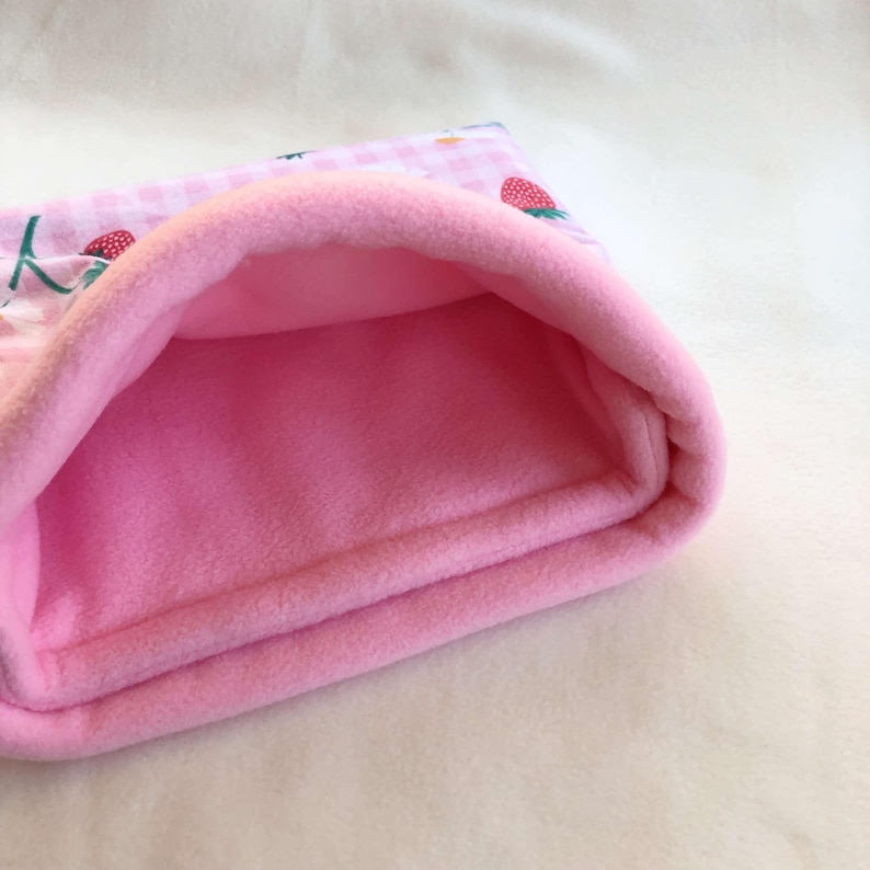 May include: A soft, plush pouch featuring a light pink and white gingham pattern on the exterior with a strawberry print. The interior is lined with a solid, bright pink fleece fabric, creating a cozy, open-ended tunnel-like structure.