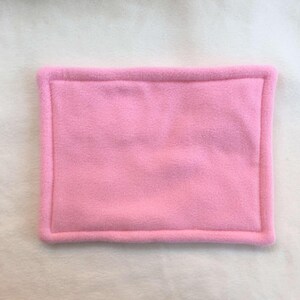 May include: A rectangular, solid light pink fleece mat featuring a soft, fuzzy texture and a thick, raised border that frames the central panel. The item is constructed from a single color of fabric with consistent stitching along the edges.