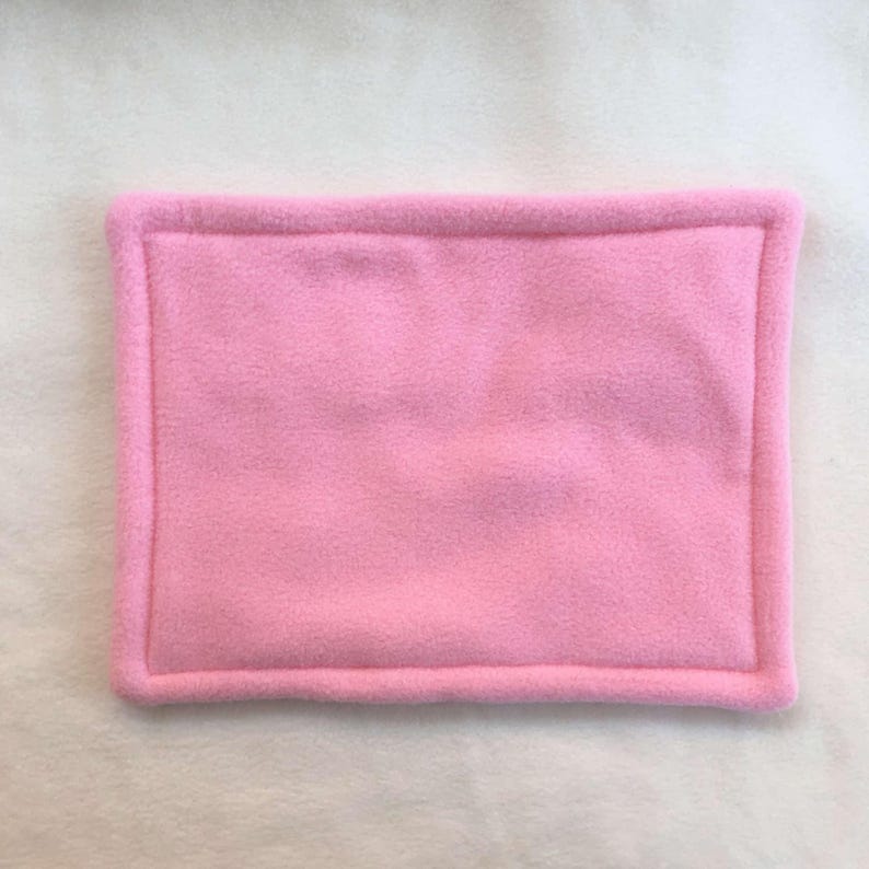 May include: A rectangular, solid light pink fleece mat featuring a soft, fuzzy texture and a thick, raised border that frames the central panel. The item is constructed from a single color of fabric with consistent stitching along the edges.