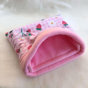 May include: A soft pet sleeping pouch featuring a light pink and white gingham pattern with a print of red strawberries and white daisies. The interior is lined with solid light pink fleece fabric, and a small black paw print tag is attached.