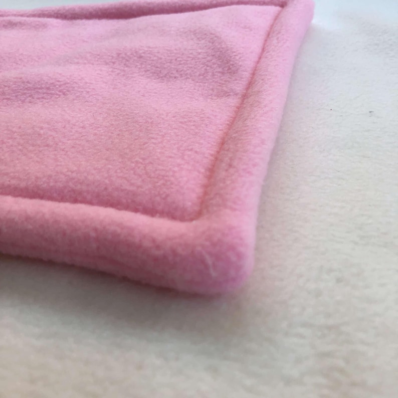 May include: A soft, light pink fleece fabric piece with a rectangular shape and a thick, raised border edge. The material has a fuzzy, textured surface throughout, showing a consistent pastel pink color and a plush, dense pile construction.