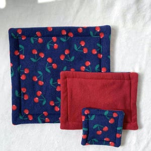 May include: A collection of three fleece pads. Two are rectangular, one is square. One rectangular pad is plain red. The other two are navy blue with a pattern of red cherries and green leaves. These pads are likely for pet bedding.