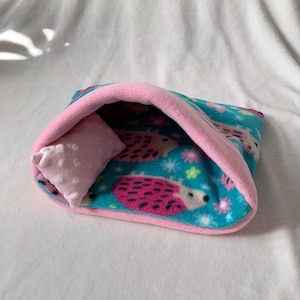 Könnte beinhalten: Ein teal- und rosa-farbenes Haustierbett mit einem passenden rosa Kissen. Das Bett besteht aus weichem Fleece-Material und zeigt ein Muster aus Igeln, Blumen und Sternen. Der Innenraum und die Verkleidung sind einfarbig rosa.