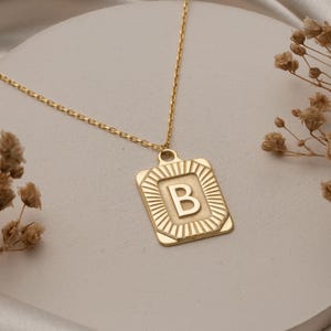 Op de afbeelding: Een goudkleurige ketting met een hanger met de letter "B" in een vierkant frame. De hanger heeft een zonnestraalontwerp rond de letter. De ketting wordt gepresenteerd op een wit, cirkelvormig oppervlak met een zachte, getextureerde achtergrond.