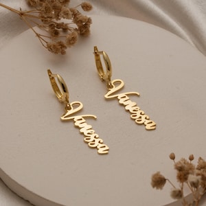 May include: Two gold-coloured hoop earrings, each with the name "Vanessa" in cursive script. The earrings are presented on a white, circular surface, with dried floral decorations. The jewellery is set against a soft, neutral background.
