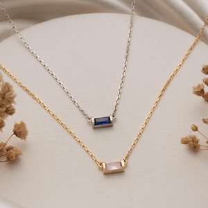 Könnte beinhalten: Zwei zarte Halsketten mit rechteckigen Anhängern. Eine Halskette ist goldfarben mit einem rosa Anhänger, die andere silberfarben mit einem blauen Anhänger. Beide Halsketten haben feine Ketten und werden auf einer weißen Oberfläche mit Trockenblumen präsentiert. Die Anhänger sind ca. 1 cm lang.
