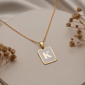 May include: A gold-tone necklace with a rectangular pendant displaying the letter "K" on a white background. The pendant is framed in gold and suspended from a fine chain. The necklace is displayed on a white surface with dried flowers.