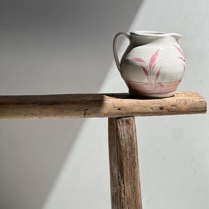 May include: A ceramic pitcher with a handle, featuring a white body with pink floral designs and a pink base. The pitcher sits on a rustic wooden bench. The background is a plain white wall with a shadow.