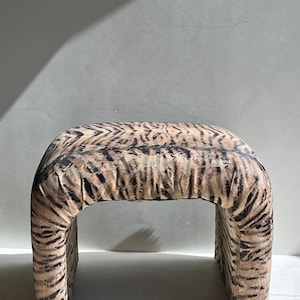 May include: A U-shaped ottoman upholstered in a textured fabric featuring a tan and black tiger stripe pattern. The piece has a continuous, curved silhouette with a flat top surface and two vertical sides that extend down to the base.