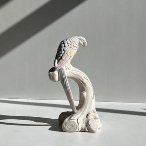 May include: A pastel-colored ceramic parrot figurine perched on a stylized tree branch. The parrot features shades of blue, pink, and yellow. The branch and base are white. The sculpture is a decorative item.