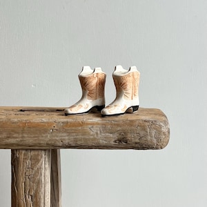 May include: A pair of ceramic cowboy boot figurines. The boots are white with tan and black accents, and are displayed on a rustic wooden surface. The boots have detailed designs and small heels.