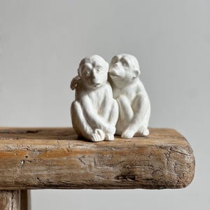 May include: A white ceramic sculpture of two monkeys in a close embrace. The figures are detailed with expressive faces and seated on a weathered wooden surface. The sculpture is a decorative item.