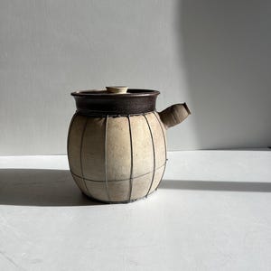 May include: A beige ceramic pot with a dark brown rim and a small lid. The pot has a rounded shape with a wire cage design. It features a spout on the side, suggesting it's for pouring. The pot is likely for kitchen or home decor.