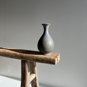 May include: A small, dark gray ceramic vase with a narrow neck and rounded body. The vase is displayed on a weathered, wooden bench with visible grain and knots. The background is a plain, light gray wall.
