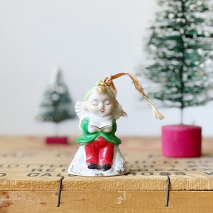 May include: Vintage porcelain Christmas ornament of a girl in a green dress and red pants singing from a book. The ornament is attached to a gold ribbon.