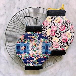 May include: Two floral print pot holders with black trim and handles. One is blue with a checkered pattern, the other is yellow, pink, and black. They are placed on a metal cooling rack.