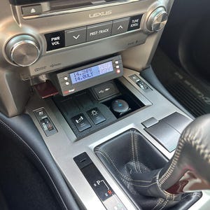 May include: Interior view of a Lexus car dashboard. The dashboard is silver and black, with a digital display showing readings. There are buttons for power, volume, and track selection. A gear shift and other controls are visible.
