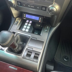 May include: A close-up of a car's dashboard with a digital display showing "329.111E 14.1ULT 75TFT 103WT". The display is mounted above a panel of buttons with a silver gear shift knob below. The dashboard is labeled "LEXUS" and has a "PWR VOL" knob, a "PRST-TRACK" button, a "TUNE SCROLL" knob, and a "SPEED PUSH 4WD" button.