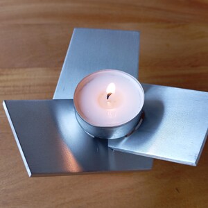 Puede incluir: Un portavelas moderno con acabado de metal plateado. El portavelas presenta tres placas rectangulares dispuestas en forma triangular, sosteniendo una vela de té encendida. La llama de la vela emite un brillo cálido.