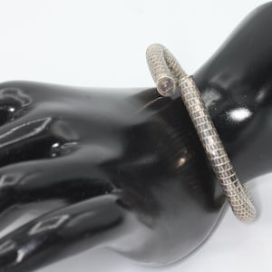 May include: A silver-toned bracelet with a flexible, segmented, grid-like texture. The bracelet features a bypass design with two rounded, flat ends. The metal surface has a metallic sheen and a repeating square pattern throughout the entire band.
