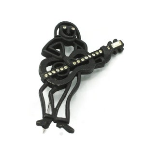 May include: Black brooch in the shape of a person playing a guitar. The brooch has a matte finish and is adorned with small, clear rhinestones. The guitar's fretboard and the person's eyes are embellished with the rhinestones. The brooch has a pin on the back.
