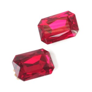 May include: Two rectangular loose gemstones with a vibrant, deep red color. Each stone features a faceted cut that creates sharp geometric patterns and light reflections across the surface, showcasing a high level of clarity and saturation.