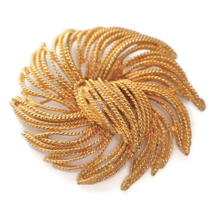 May include: A gold-toned brooch with a textured, woven design. The brooch features a spiral pattern with radiating lines, creating a floral or sunburst effect. The jewelry piece is set against a white background.