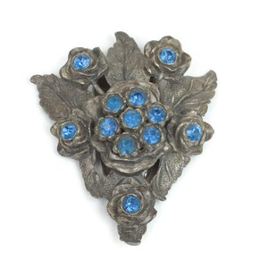 May include: A vintage silver-toned brooch in a floral design. The brooch features several rose-shaped elements and leaves, with bright blue gemstones set in the center of the roses. The brooch is set against a white background.