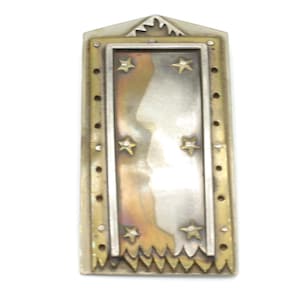 May include: A rectangular metal brooch featuring a central panel with a gradient from dark bronze to silver, decorated with six small gold-toned stars. The frame is brass-colored with a series of small circular indentations and a jagged bottom edge.