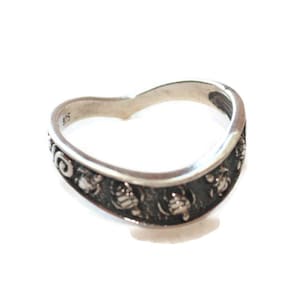 May include: A silver ring with a curved design. The ring features a band with a series of small, detailed floral or ornamental patterns. The ring is set against a white background.