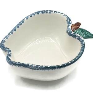 May include: White ceramic apple-shaped bowl with a blue rim and a green leaf and brown stem detail. The bowl has a smooth interior and a slightly textured rim, perfect for serving snacks or small dishes.