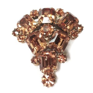 May include: A vintage brooch featuring a cluster of faceted, amber-colored rhinestones. The brooch has a symmetrical design with larger rectangular stones at the center and smaller, round stones around the edges. The metal setting is a muted gold tone.