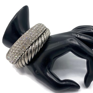 May include: A silver-toned bracelet with a row of sparkling rhinestones and a twisted rope design. The bracelet is displayed on a black hand-shaped jewelry holder, showcasing its elegant and glamorous style. This accessory is perfect for adding a touch of sparkle to any outfit.