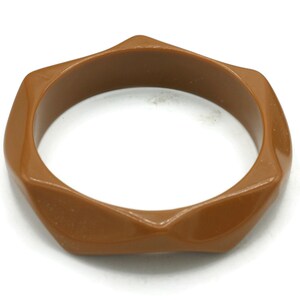 May include: A solid brown bangle bracelet with a smooth, glossy finish. The bracelet features a distinct geometric, hexagonal-inspired shape with six prominent, angled points along the outer edge, creating a structured and uniform appearance.